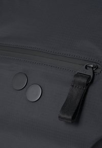 Detail einer schwarzen Stofftasche mit einem Reißverschlussfach, zwei runden schwarzen Knöpfen und einem strukturierten Riemen zum Schließen oder Verstellen.