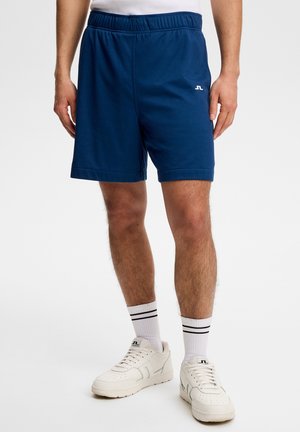 Mand iført navyblå atletikshorts, hvide sneakers og hvide sokker med sorte striber, stående på ensfarvet hvid baggrund.