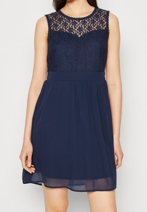 Woman wearing sleeveless navy dress with floral lace bodice and sheer overlay skirt against a plain light background.