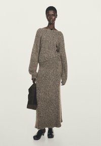 Gehaakte, beige en bruine trui en rokset met lange mouwen, een relaxte pasvorm en textuurstof. Gepaard met een zwarte tas en klompen.