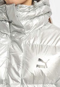 Nærbillede af en sølvfarvet oppustelig jakke med høj krave og Puma-logo på brystet, båret af en person med langt brunt hår.