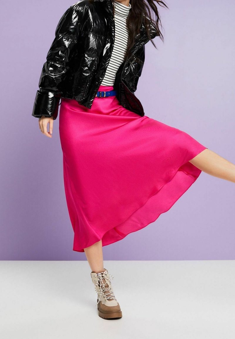 Schwarze, glänzende Steppjacke über einem schwarz-weißen gestreiften Oberteil, kombiniert mit einem fließenden, rosa Satinrock und beigen Plateau-Stiefeln mit Schnürung.