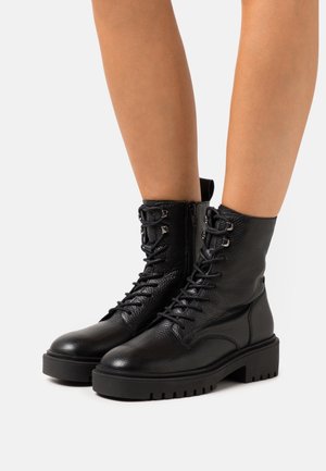 Black leather lace-up ankle boots with thick soles worn on bare legs against a plain white background.