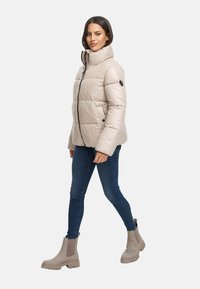Giacca puffer beige con colletto alto e zip anteriore, abbinata a jeans blu scuro e stivaletti beige. Design strutturato e trapuntato.