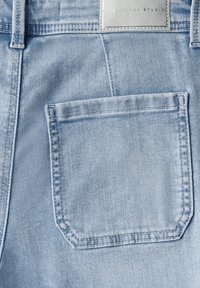 Jean en denim bleu clair avec une poche arrière et une étiquette en simili cuir positionnée au-dessus. Le tissu a une texture lisse avec des coutures visibles.