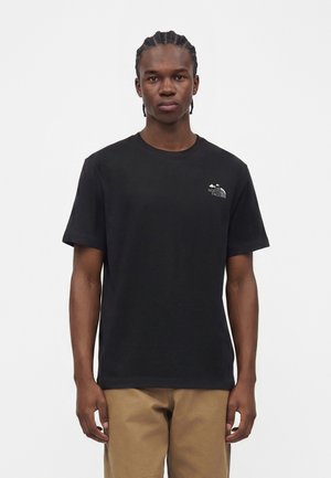 Junger Mann mit geflochtenem Haar, der ein schwarzes The North Face T-Shirt und beige Hose trägt, steht vor einem einfachen weißen Hintergrund.