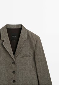 Rusinapahvan houndstooth-takkia, jossa on lovettu kaulus, kolme mustaa nappia ja rintatasku; pehmeä rakenne, räätälöity istuvuus ja minimalistinen muotoilu.