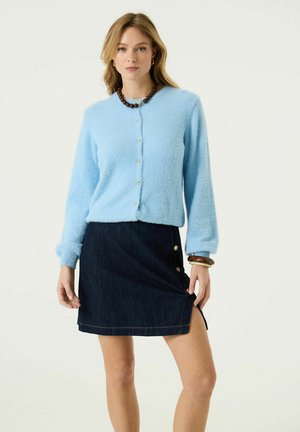 Frau trägt hellblauen flauschigen Cardigan mit Knöpfen und dunkelblauen Denimrock mit seitlichen Knöpfen und Schlitz, accessoriert mit Halskette und Armbändern.