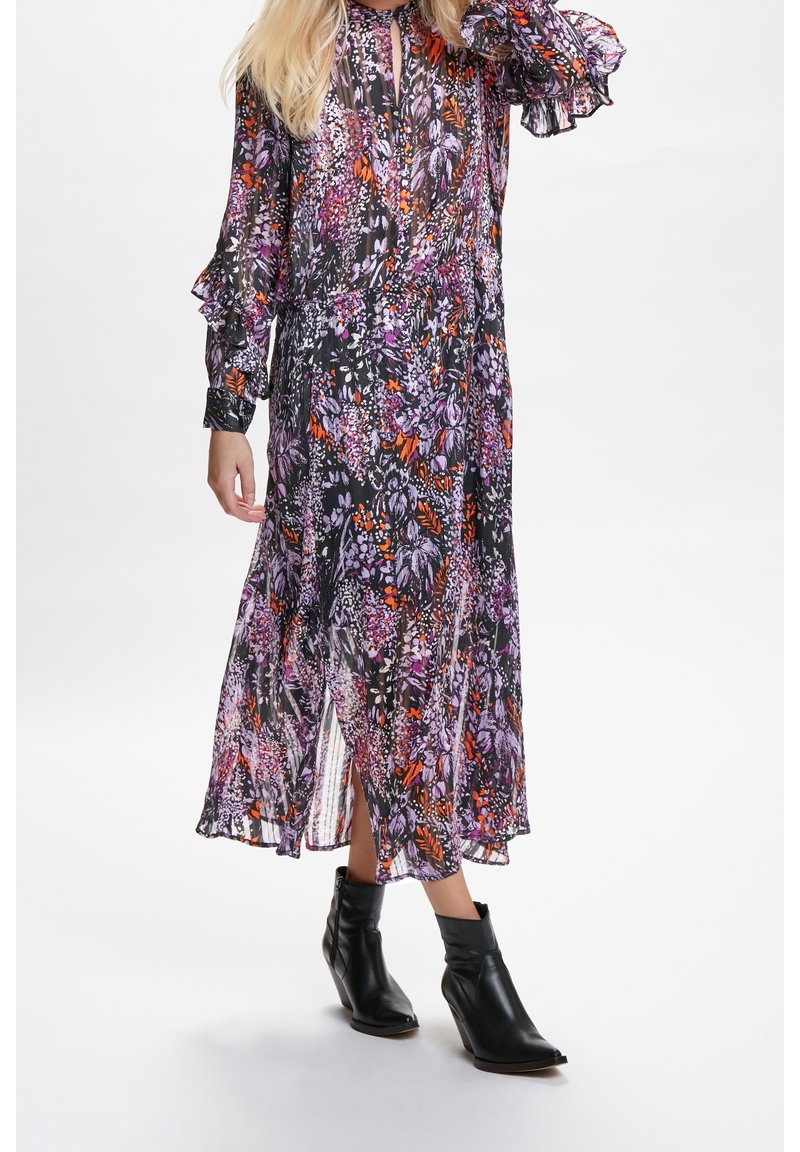 Femme portant une robe à manches longues à fleurs dans des tons violet et orange, associée à des bottines noires sur un fond uni.