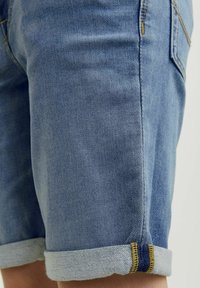 Vaaleansiniset farkkushortsit, joissa on käänteet. Ominaisuuksiin kuuluu keltainen ompelu ja kontrastivärinen tummansininen korostus lahkeessa. Sujuva pinta, rento muotoilu.