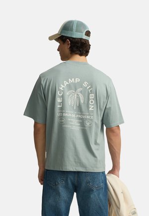 Hombre con camiseta azul verdosa clara con gráfico de palmera y texto en la espalda, vaqueros azules, gorra de béisbol verde de malla, sosteniendo chaqueta beige.