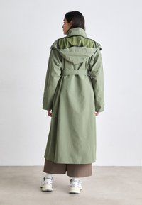 Trench coat verde lungo con vita aderente, cappuccio e colore interno a contrasto. Presenta un design con cintura e maniche lunghe.