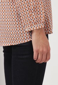 Camicia a maniche lunghe con un motivo a rombi arancioni e bianchi, caratterizzata da un dettaglio ai polsini con bottoni e un tessuto leggero e testurizzato.