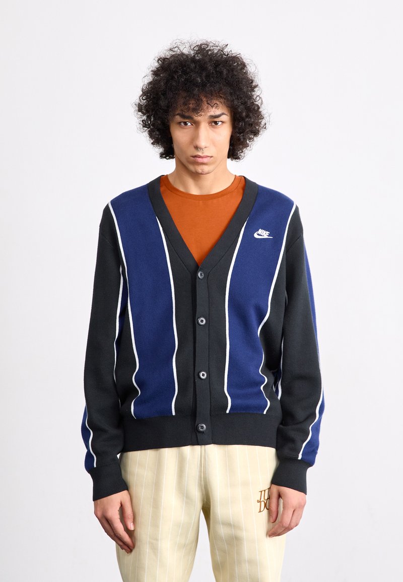 Zwarte en marineblauwe cardigan met witte strepen, v-hals, knoopsluiting en Nike-logo. Draagt over een oranje shirt, gecombineerd met gestreepte broek.