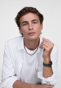 Un giovane in camicia bianca con colletto sopra una maglietta bianca, indossa una collana nera e un orologio in rete con quadrante circolare scuro.