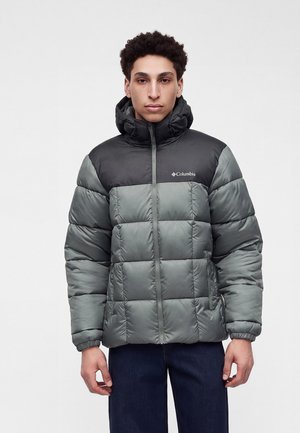 Jeune homme portant une doudoune gris et noire Columbia avec capuche, debout les bras détendus sur un fond clair uni.