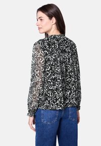 Durchsichtige schwarze Bluse mit weißem Blumenmuster, langen Ärmeln und gerafftem Rückendetail. Kombiniert mit Jeans.