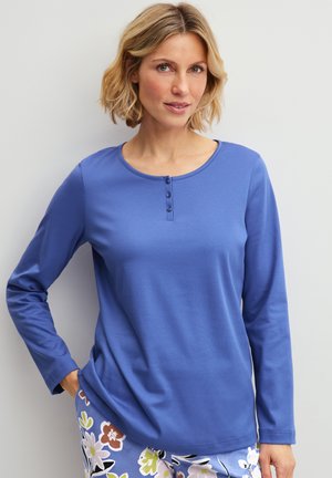 Frau mit kurzen blonden Haaren, die ein blaues Henley-Shirt mit langen Ärmeln und eine Hose mit Blumenmuster trägt, steht vor einem einfarbigen hellen Hintergrund.