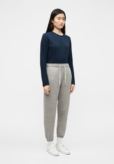 Ασιάτισσα γυναίκα όρθια, φοράει μπλε μακρυμάνικο πουκάμισο, γκρι παντελόνι φόρμας με κορδόνι,  λευκές κάλτσες και λευκά αθλητικά, με φόντο απλό λευκό τοίχο.