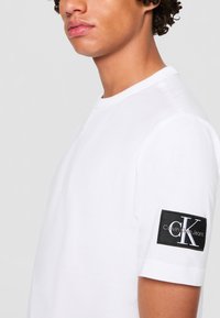 Persona che indossa una camicia bianca a maniche corte con una toppa nera con il logo Calvin Klein Jeans sulla manica sinistra, ritagliata al mento e alla spalla.
