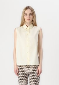 Blusa de manga corta, de color amarillo claro, con cuello y cierre de botones en la parte delantera. Combinada con pantalones ajustados y estampados en colores negro, beige y blanco.