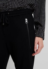 Pantalones de chándal negros con tejido texturizado, que presentan una cintura ajustable, un bolsillo lateral con cremallera y herrajes plateados, combinados con una camiseta negra de estampado.
