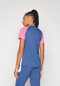 Μπλε αθλητικό πουκάμισο με ροζ μανίκια, κατασκευασμένο από διαπνέον ύφασμα. Διαθέτει λευκές πλαϊνές λεπτομέρειες και αθλητικό, εφαρμοστό σχέδιο.