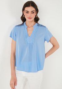 Blusa blu chiaro con maniche corte, caratterizzata da un scollo a V, dettagli in pizzo sulle spalle e una vestibilità ampia e fluida.