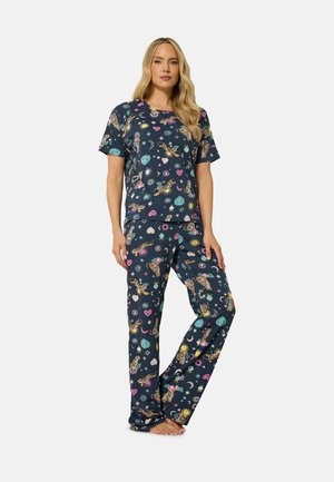 Kvinde står barfodet iført matchende mørkeblå pyjamas med farverige tiger-, sol-, måne- og hjerteprint på en ensfarvet baggrund.