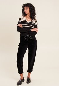 Gestreifter V-Ausschnitt-Pullover in Beige und Schwarz, kombiniert mit schwarzen Cordhosen mit einem Kordelzug. Schwarze Slipper runden den Look ab.