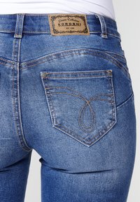 Jean en denim bleu au design ajusté. Détails de couture marquants et un motif décoratif sur la poche arrière, avec une étiquette en cuir à la taille.