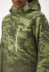 Zelena jakna s mramornim tamnozelenim uzorkom, s naglašenim kapuljačom i žutim patentnim zatvaračem. Oakley logo prikazan na prednjoj strani.