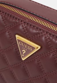 Burgunderfärgad faux läderklutch med kviltat mönster, guldfärgad dragkedja och triangulär guldvarumärkessegen med texten "GUESS U.S.A."