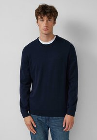 Giovane uomo con capelli castani ricci che indossa un maglione girocollo blu navy su una camicia bianca e jeans blu, in piedi davanti a un muro grigio.