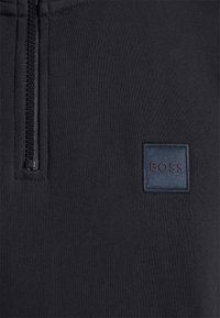 Svart sweatshirt med halv dragkedja, med en texturerad marinblå logotypmärke med "BOSS" på vänster sida. Mjukt tyg och avslappnad design.