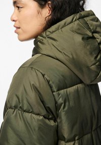 Olīvu zaļa pūkaina jaka ar kapuci, ar quiltēta dizaina, gludu tekstūru un vieglu spīdumu. Redzamās šuves un plecu vīles.