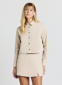 Maglietta beige a coste con colletto, bottoni sul davanti e maniche lunghe, abbinata a una gonna corta coordinata con spacchi laterali.