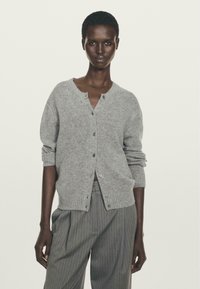 Donna nera con capelli corti indossa un cardigan di maglina grigio con bottoni e pantaloni a vita alta grigio a righe, in piedi contro uno sfondo neutro.