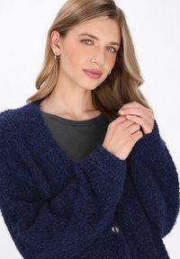 Marineblauer, flauschiger Cardigan mit tiefem V-Ausschnitt und strukturiertem Stoff. Lange Ärmel und eine Knopfdetail vorne. Lockere Passform.