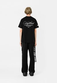 Camiseta de algodón negra con texto en blanco en la parte posterior. Combinada con pantalones negros holgados que muestran detalles gráficos y bolsillos laterales.