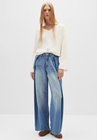 Maglione a coste beige chiaro con scollo a V, abbinato a jeans in denim blu sbiadito a gamba larga. Presenta una vita a media altezza e una linea dritta.