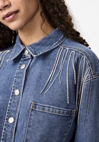 Denimist särgid hõbedaste rhinestone-aktsentidega, mis voolavad õlgadelt, millel on krae, esitusega nupud ja rinna tasku.