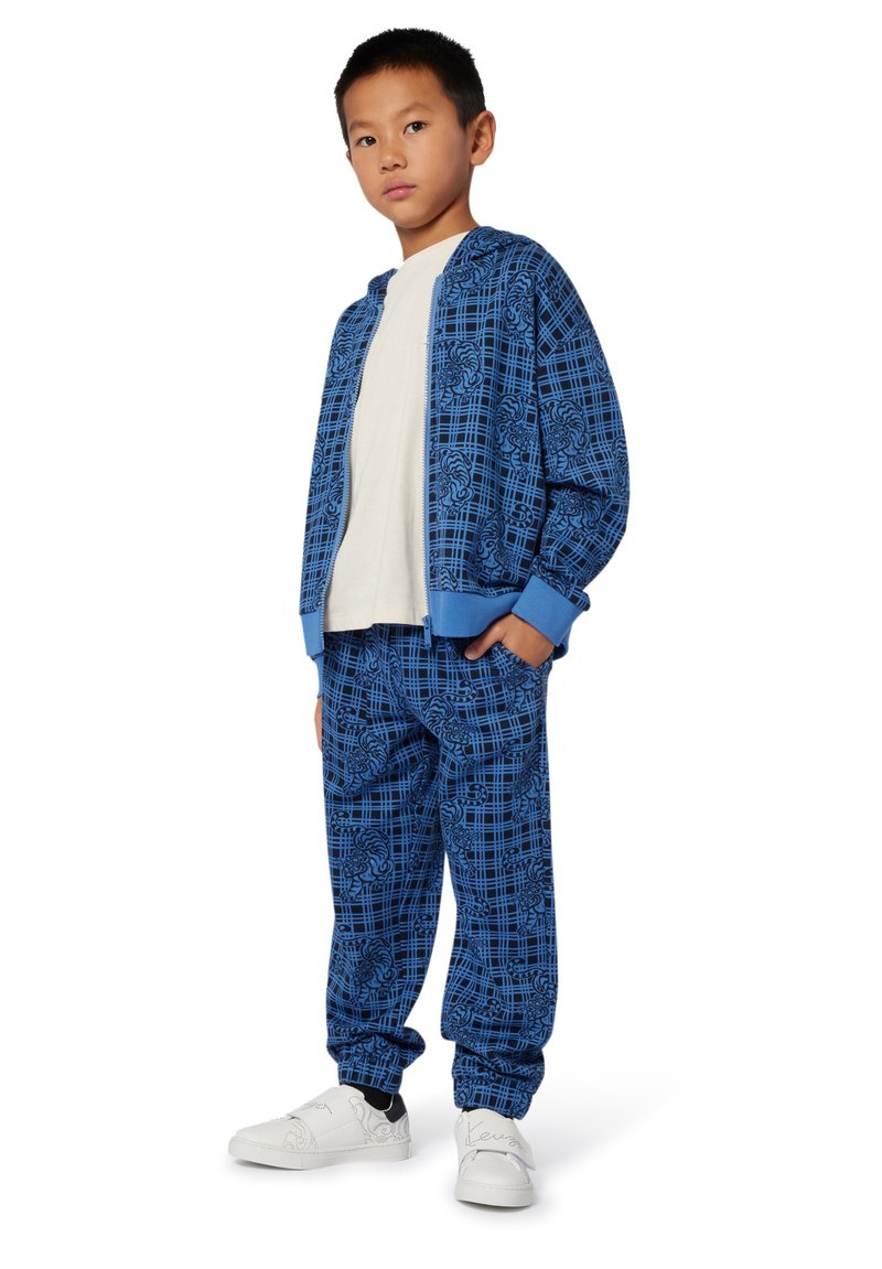 Survêtement bleu avec un motif géométrique, comprenant une veste à fermeture éclair et un pantalon à taille élastique. Porté avec des chaussures blanches.