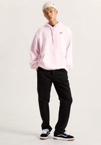 Rosa Kapuzensweatshirt mit einer Vorderentasche, kombiniert mit schwarzen Hosen und schwarzen Sneakers. Das Sweatshirt hat ein kleines Logo auf der Brust.