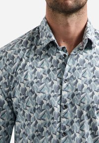 Man in een dichtgeknoopte overhemd met een blauw en grijs abstract geometrisch patroon, waarbij details van de nek en onderkant van het gezicht zichtbaar zijn.