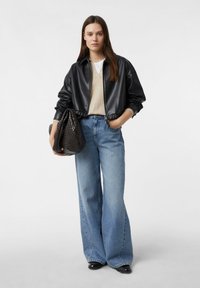 Czarna kurtka typu bomber z czarnej skóry ekologicznej, beżowy top, jasnoniebieskie dżinsy o szerokich nogawkach, trzymająca czarną plecioną torbę. Prosty krój, luźne dopasowanie i gładka faktura.