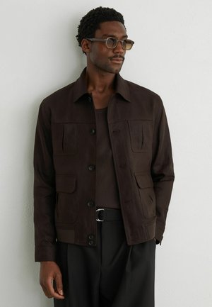 Mand iført mørkebrunt knappet jakke, brun striktrøje, sorte bukser med sølvspænde, og ravfarvede rektangulære solbriller, stående mod en hvid væg.