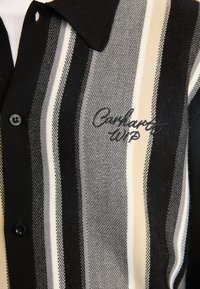 Camicia a righe nera e grigia con colletto, dotata di chiusura a bottone e logo ricamato "Carhartt WIP". Tessuto lavorato a maglia.