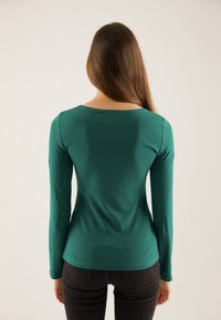 Long-sleeve teal top in a soft fabric with a round neckline and fitted design, paired with dark jeans, viewed from the back.