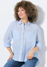 Hellblaue gestreifte Hemdbluse mit zwei Fronttaschen, aufgerollten Ärmeln und einem etwas längeren Rückensaum. Hergestellt aus einem weichen, leichten Stoff.